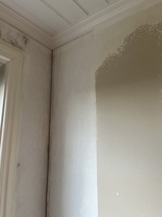 Gipshörn med delvis avlägsnad tapet, vitt papperstoftigt rest kvar på väggen, synliga kanter och ett fönster bredvid.