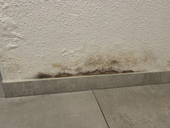 Fuktskadad vägg med avflagad färg och missfärgningar nära golvet i 60-tals villa efter vattenläcka, omgiven av klinkergolv.