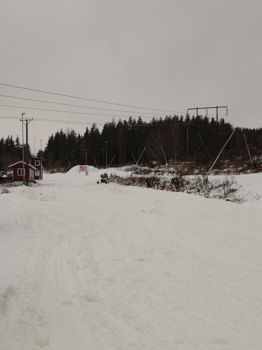 Snötäckt landskap med en röd byggnad till vänster och en skog i bakgrunden. Elektriska ledningar korsar himlen över snön.