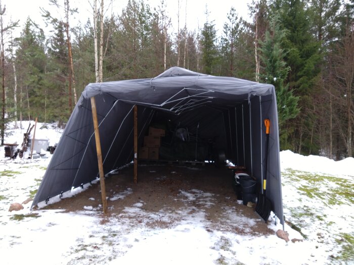 Garagetält med böjda sidostolpar och knäckta takstolar under snötäckt mark, omgiven av skog.