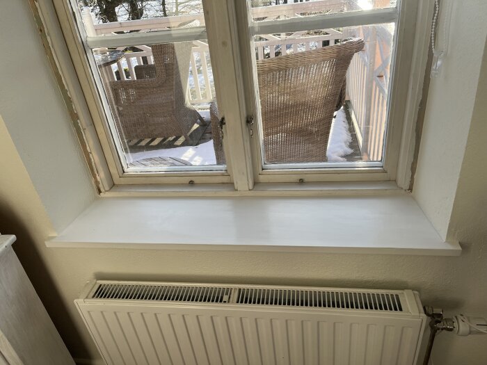 Fönsterbräda vid ett fönster, eventuellt nyss silikonad, med utsikt mot en snötäckt altan med flätade möbler.
