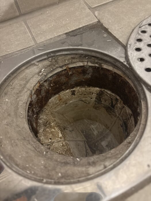Öppen golvbrunn i badrumsgolv med rost och smuts, saknar tätning. Synlig betong runt öppningen där dusch ska installeras.