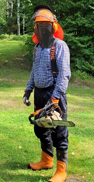 Person med skyddsutrustning inklusive hjälm och skyddsbyxor, håller en motorsåg i en skogsmiljö.