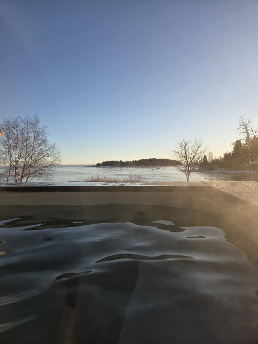 Vy från ett utomhusvarmbad med ångande vatten under en klar winterhimmel, omgiven av snöiga stränder och träd nära en stilla sjö.