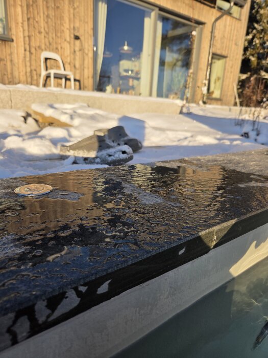 Uteplats med snötäckt mark och en träbyggnad i bakgrunden, sedd från kanten av ett varmbad med vatten.