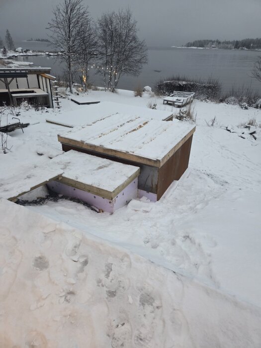Snötäckt trädgård med provisoriskt lock av cellplast över ett varmbad, beläget vid en frusen sjö, omgiven av snötäckta träd och bänkar.