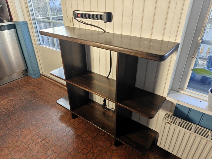 Nybyggd svartbrun hylla med tre nivåer i köket, avsedd för kaffekokare och utrustning, med eluttag monterat på väggen bakom.