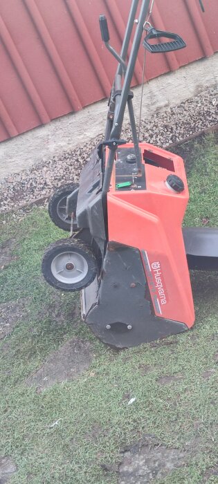 Röd snöslunga av märket Husqvarna står lutad mot en vägg, fotograferad ute på en gräsmatta.