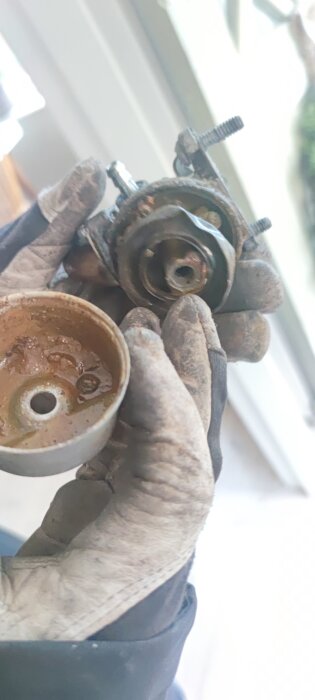 En nedmonterad förgasare från en slunga, visas tillsammans med en skålformad del, hålls av en person i arbetshandskar.