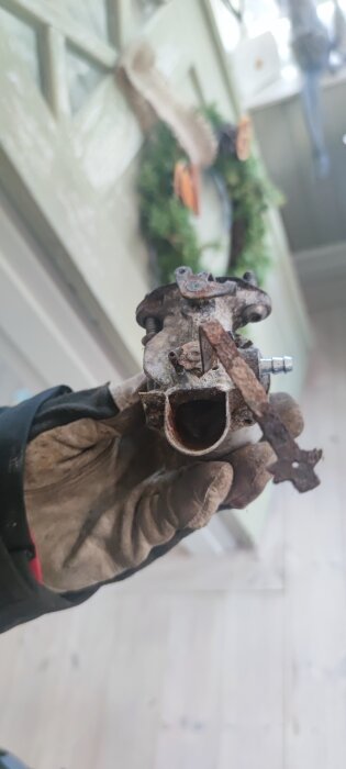 Rostig förgasare till snöslunga hålls i handskbeklädd hand, med bakgrund av diffus inomhusmiljö.