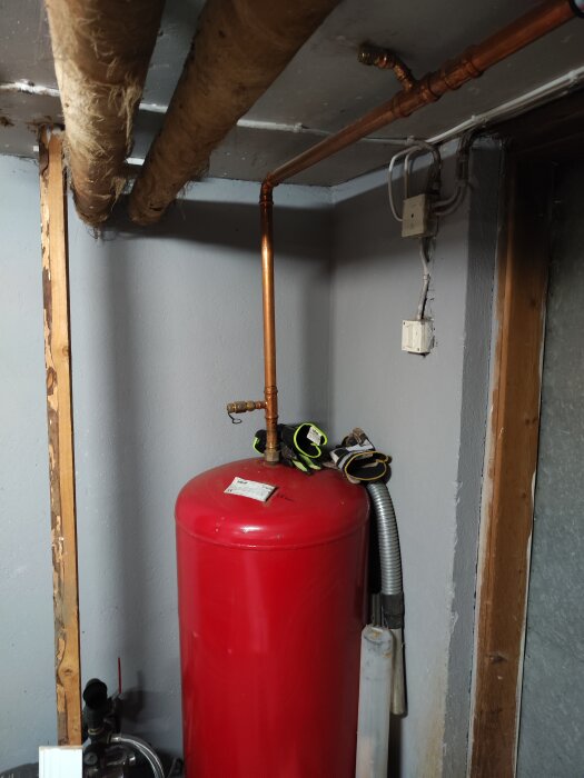 Rött expansionskärl i källare med kopparrör anslutna ovanifrån, samt skyddshandskar vilandes ovanpå kärlet.