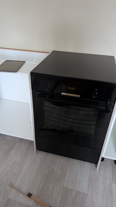 Svart ugn och vita köksskåp under installation; golvet syns med en träbit i bildens nederkant.