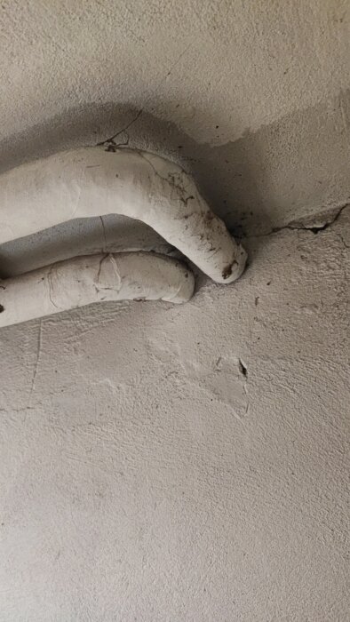 Två böjda rör täckta av vit isolering monterade mot en ojämn, krackelerad vägg i en källare.