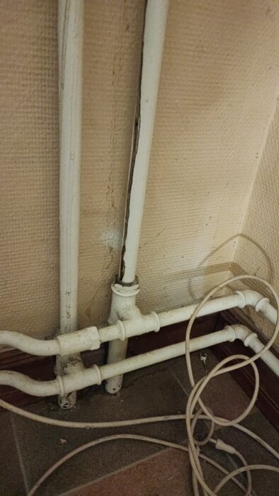 Rörsystem i källare med flera kopplingar och rördragningar längs väggen och golvet, omgivet av elkablar och smutsig bakgrund.