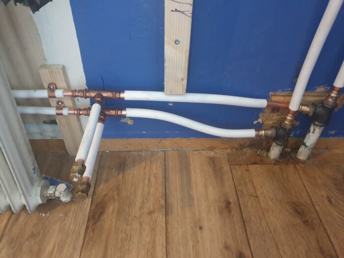 Rörkopplingar och radiatorrör i en källare mot en blå vägg, med synlig slityta på golvet och en träregel fastsatt på väggen.