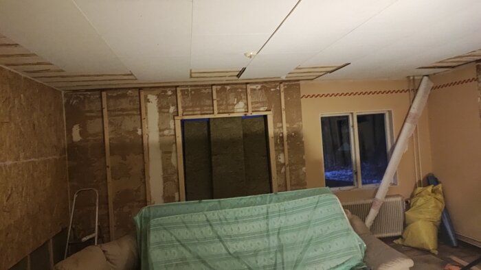 Vägg med isolerade reglar och borttaget fönster i ett rum under renovering, med en grön presenning på soffan.