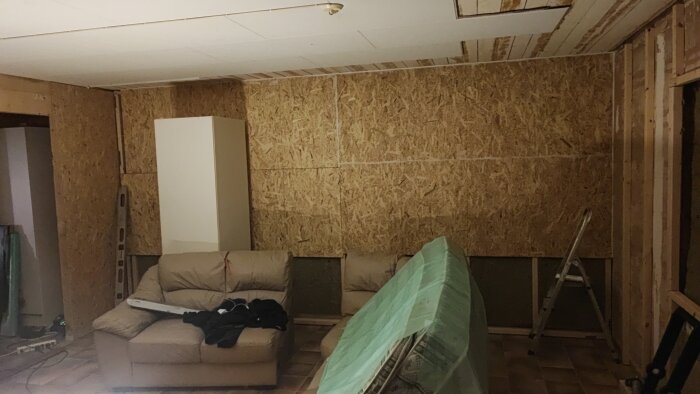 Renoveringsprojekt med osb-skivor på väggen, en soffa, en stege och skyddstäckta möbler i rummet.