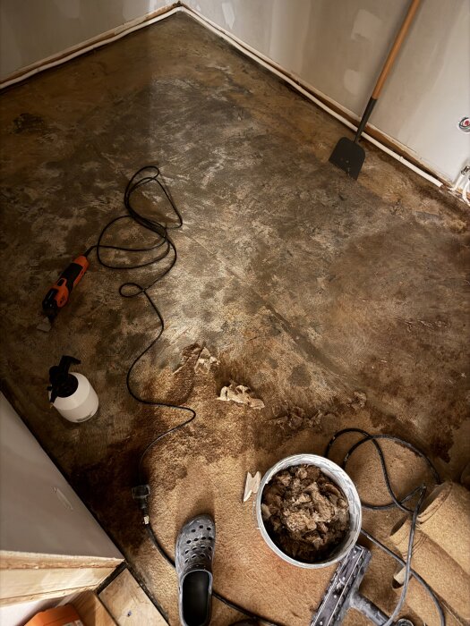 Golv med verktyg för renovering, barkjärn och skrapor synliga, samt en burk med skräprester och en sprayflaska. Ojämn yta antyder arbete pågår.