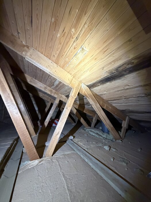 Trätakstolar och isolering på en vind med synliga träpaneler och brädor av olika dimensioner under taket.
