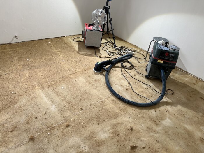 Ett rum med slipad spånskiva på golvet, en slipmaskin, sladdar och en fläkt i hörnet. Förberedelser för foam och parkettläggning.