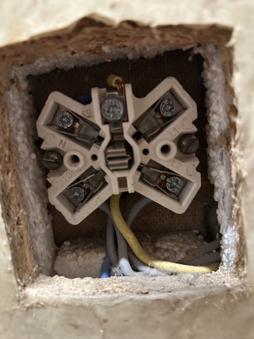 Närbild på elektriskt vägguttag med ledningar synliga, visar kopplingsschema med kablar i olika färger och flera skruvanslutningar.