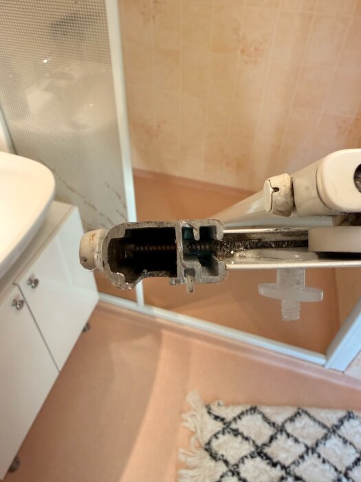 Duschväggshjul med sprucken vit plastdel, del av skjutdörrsmekanism i badrumsomgivning.