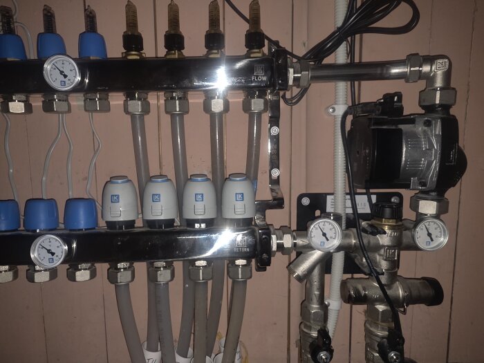 Golvvärmefördelare med termometrar och rör i ett värmesystem, installerat mot en orange vägg.