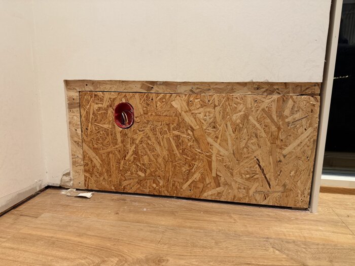 En vit vägg med OSB-skiva monterad och ett runt hål med rör synligt, troligen efter vattenläcka. Trägolv framför väggen.