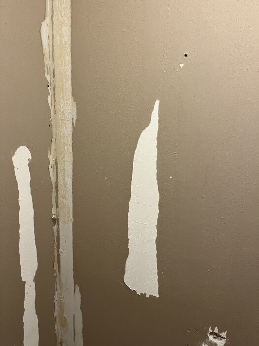 Bilden visar en brun gipsskiva med rester av plastmatta och synlig vit yta efter borttagen tejp eller lim. Skruvhål och ojämnheter syns.