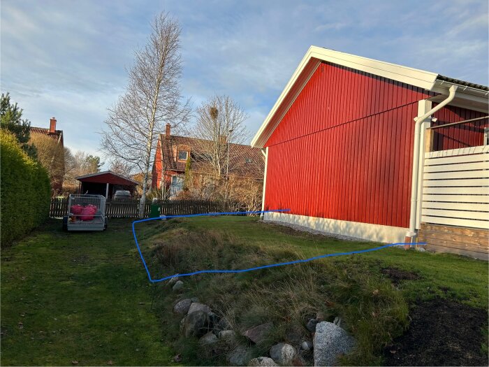 Bild på en enplansvilla i röd fasad med en blåmarkerad yta som visar planerad tillbyggnad på gräsmatta med slänt.