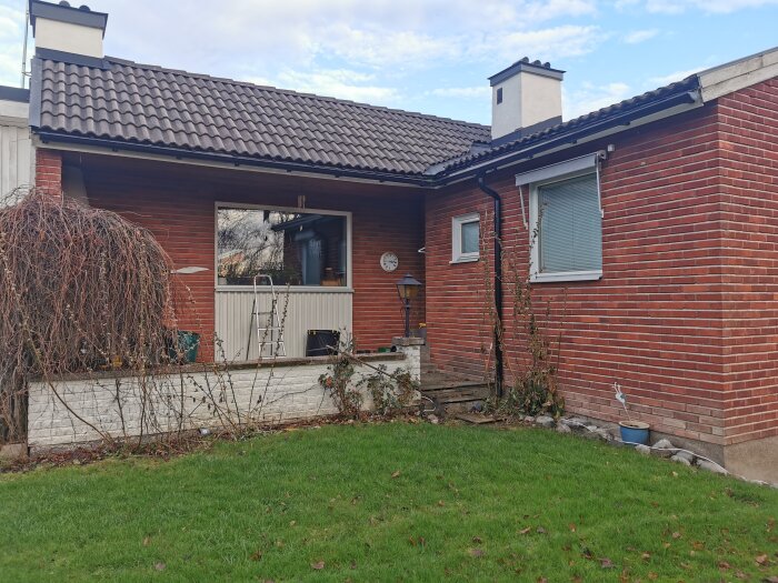 Röd tegelvägg med träpanel, vit mur, vildvuxen växt, öppna spisens skorsten visar sig. Metallräcke med vildvin framför grön gräsmatta.