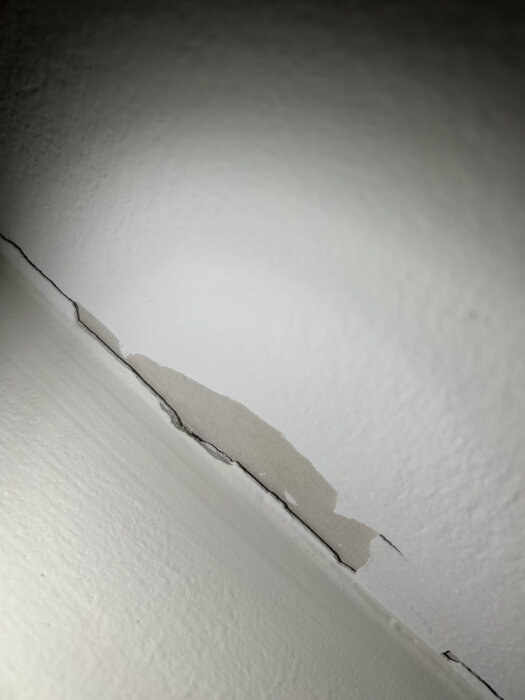 Flagnande färg och sprickor i övergången mellan vägg och tak, med blottad yta, i ett rum med äldre målning.