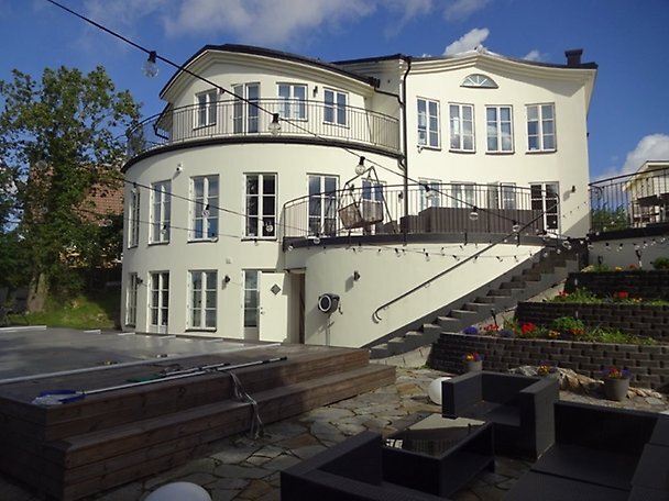 Vit arkitektritad villa i Stockholmsområdet med runda former och flera fönster. Stenlagd uteplats i förgrunden med utemöbler och poolområde.