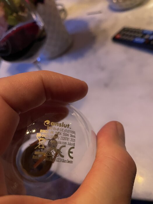 Hand håller i en glödlampa från "enslut" med text och specifikationer på ytan. Bakgrund med ett glas vin och en fjärrkontroll.