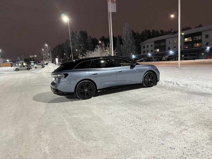 Silvergrå bil parkerad på en snöig parkeringsplats under gatubelysning, omgiven av snö och skog i bakgrunden.