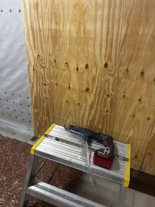 Skruvdragare med skruvband och batteri placerat på en metallstege framför en träpanelvägg.