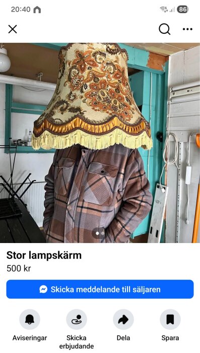Person i rutmönstrad jacka bär en stor, mönstrad lampskärm över huvudet i ett rum med teal och vita väggar. Lampan är beige med fransar.