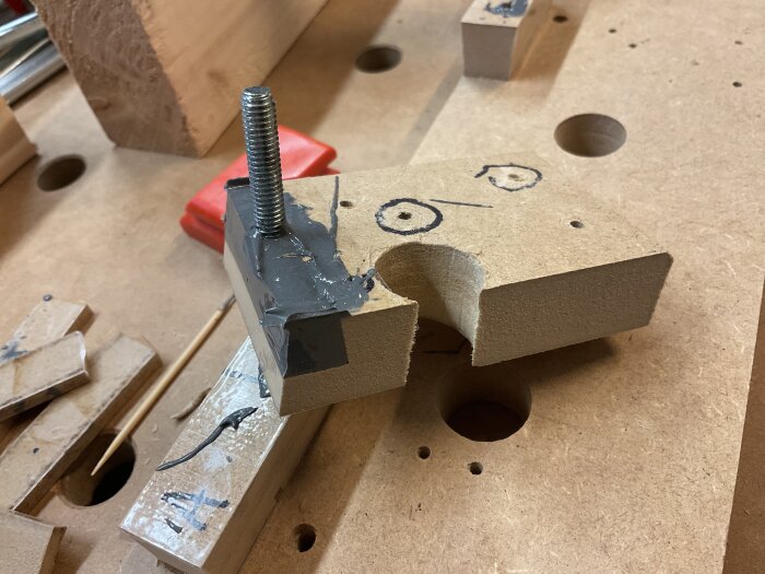 Träblock och skruv med epoxi, del av gjutprojekt, placerat på arbetsbänk med bearbetade ytor och markerade konturer för montering.