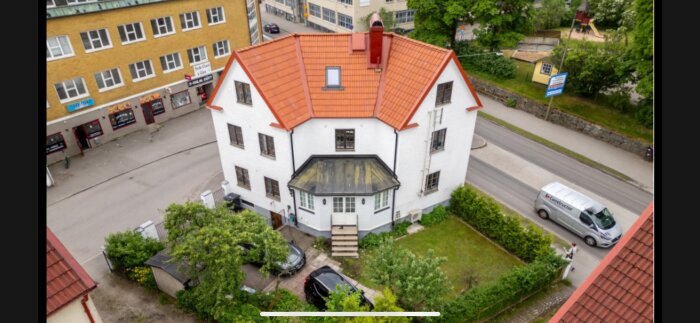 Vit byggnad med rött tegeltak, trädgård och två bilar utanför, omgiven av trottoarer och fler byggnader i ett stadskvarter.