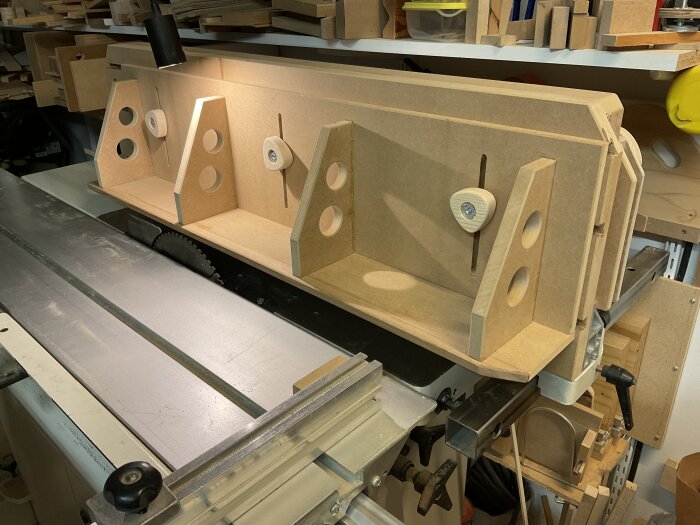 Justersåg med monterat högt anhåll och L-anhåll, omgiven av träbitar och verktyg i verkstadsmiljö.