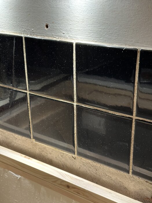 Svarta 15x15 cm kakelplattor på en vägg, med viss skada och en synlig borrhål ovanför.