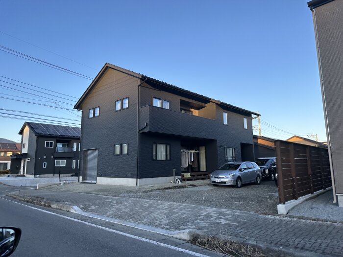 Nybyggt hus i Japan med mörk fasad och stora fönster. Två bilar parkerade framför, soliga kläder hänger på balkongen.