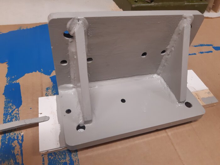 Gråmålad, svetsad metallvinkel med flera hål, monterad på en platt underdel med kartong och blå bakgrund.