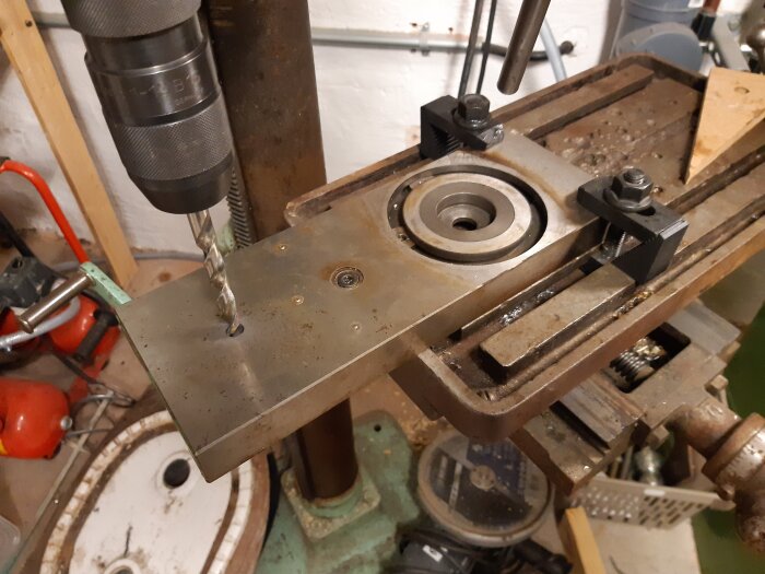 Borrning i metallstycke monterat på en svarv omvandlad till fräs, med borrspindel och metallklämmor synliga i bakgrunden.
