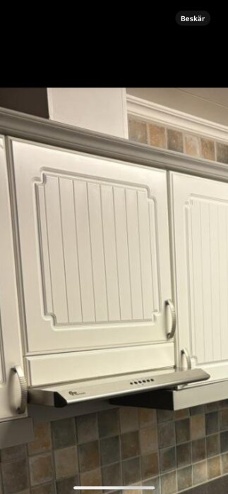 Vita köksluckor med profilerad design ovanför en rostfri köksfläkt mot en kaklad vägg.