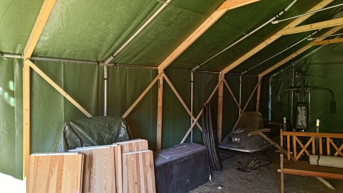 Insidan av ett stort tält med uppstöttade trästolar och metallrör, en skoter och träningsutrustning i bakgrunden.