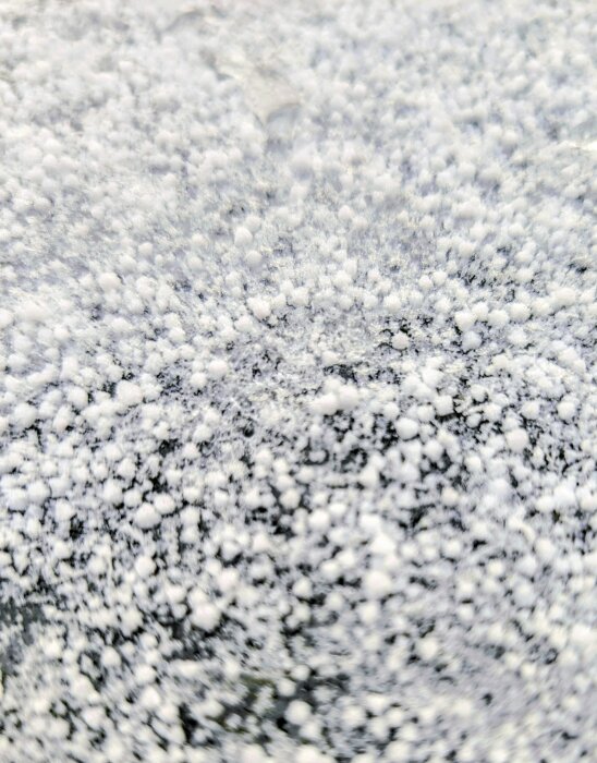 Närbild av snö eller frost på en mörk yta, täckt av små vita ispartiklar och kristaller.