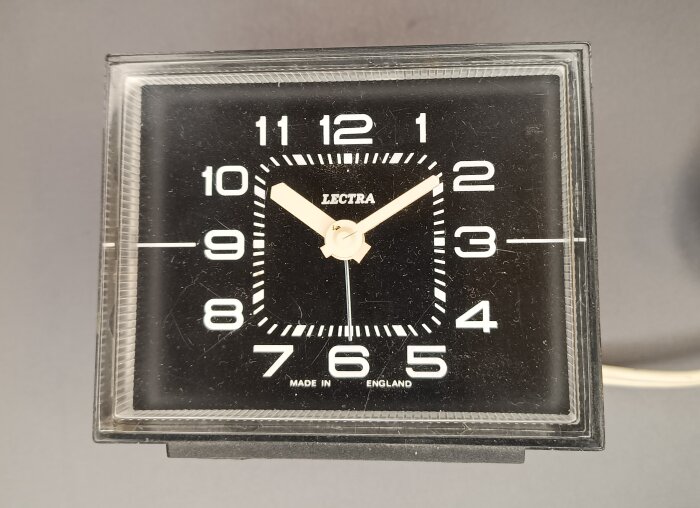 Svart synkronur med vita siffror och visare, märket "LECTRA" syns på urtavlan, beroende av 50 Hz frekvens för korrekt tidhållning.