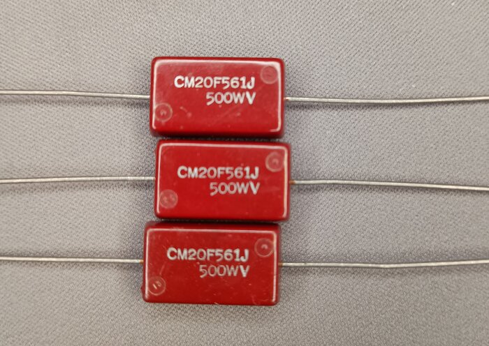 Tre röda micakondensatorer med märkningen "CM20F561J 500WV" ligger på rad med långa anslutningsben.