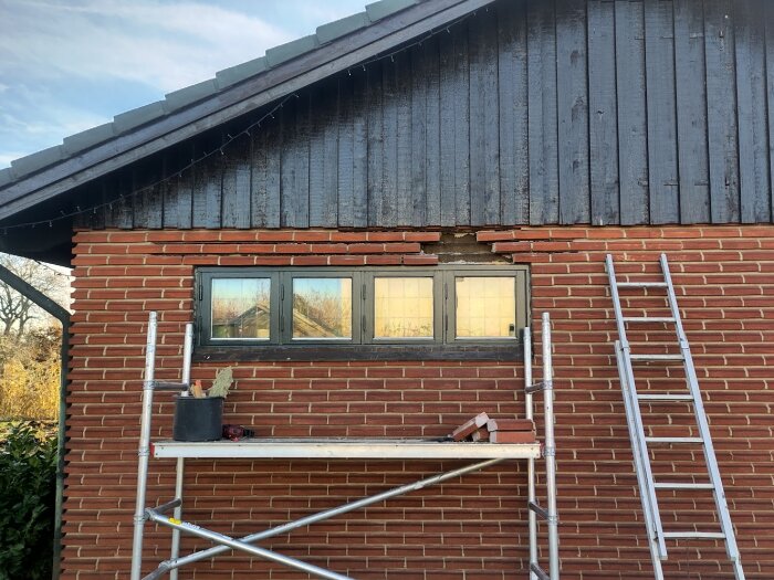 Byggarbetsplats med tegelfasad där ett stort fönster är borttaget, synliga skador i muren och byggställning med verktyg och stege.